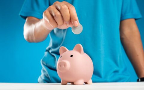 A close-up of an adult's hand dropping a coin into a piggy bank, symbolizing savings and investment.