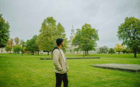A man stands in a park with a historic building and clock tower in the background, surrounded by greenery.