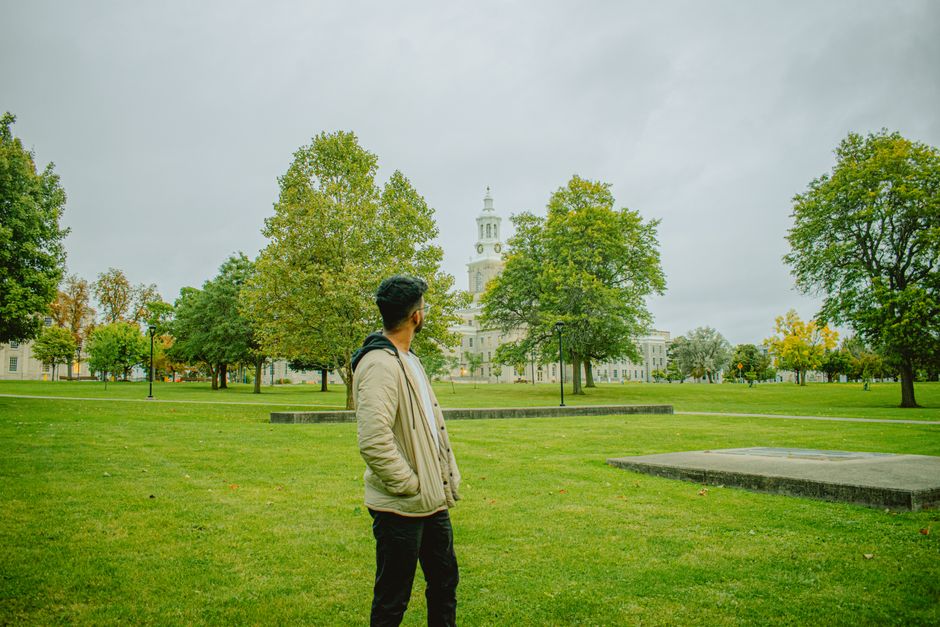 A man stands in a park with a historic building and clock tower in the background, surrounded by greenery.