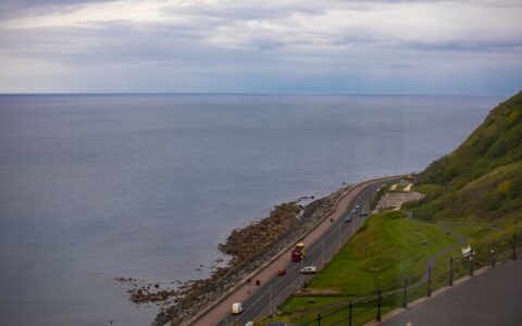 A serene coastal road view with a vast calm sea and lush green hills under a cloudy sky.