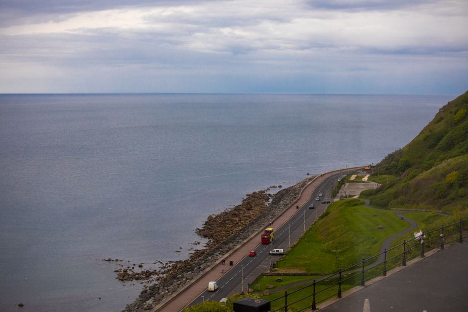 A serene coastal road view with a vast calm sea and lush green hills under a cloudy sky.