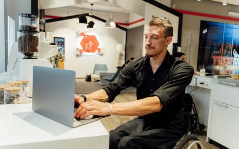 Man in a wheelchair using a laptop in a modern cafe, showcasing accessibility.