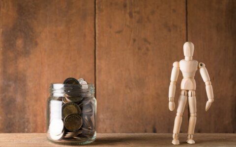 Wooden mannequin standing next to a jar filled with coins against a wooden backdrop, symbolizing savings and planning.