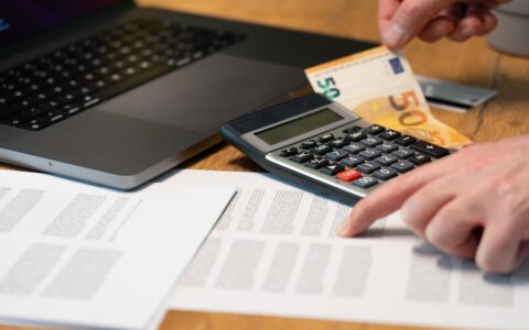 Close-up of hands using calculator with euro notes and laptop on desk.