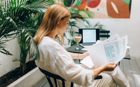 A woman reviews financial documents in an indoor garden space, with a laptop open.