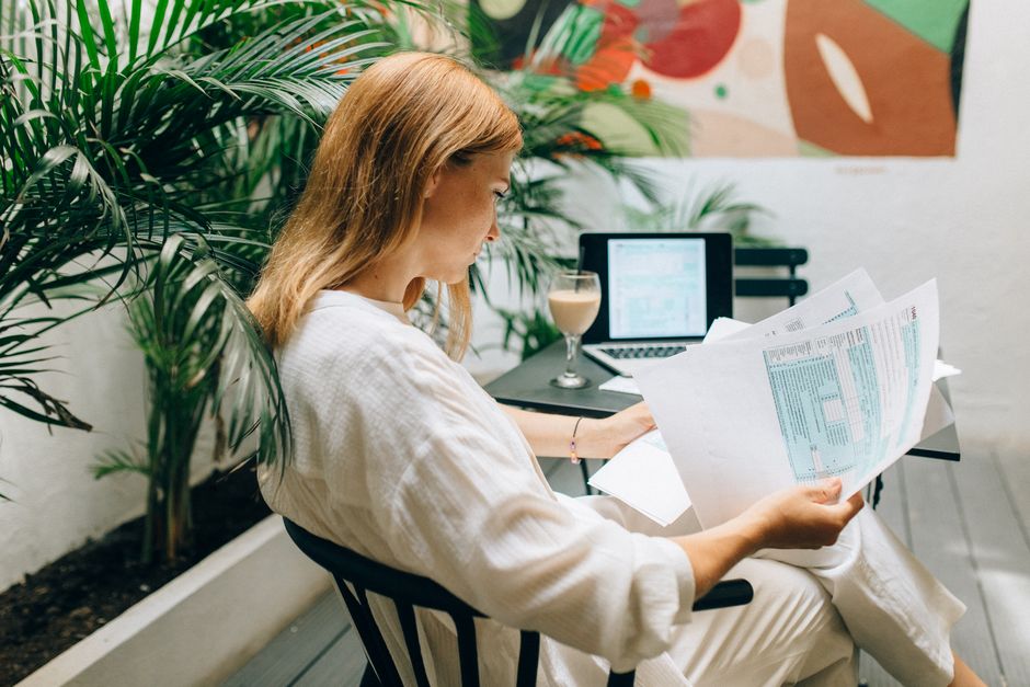 A woman reviews financial documents in an indoor garden space, with a laptop open.