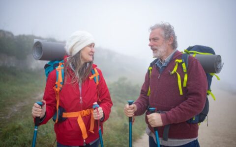 A senior couple hiking with trekking poles amidst foggy mountain landscape in Portugal, sharing a joyful moment.