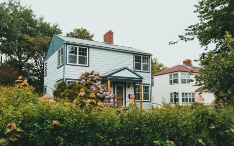 Charming two-story wooden house surrounded by lush greenery and vibrant flowers in a suburban neighborhood.