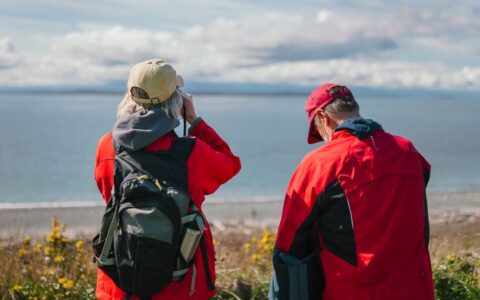 Elderly couple in red jackets enjoying seaside view, outdoors with backpacks.