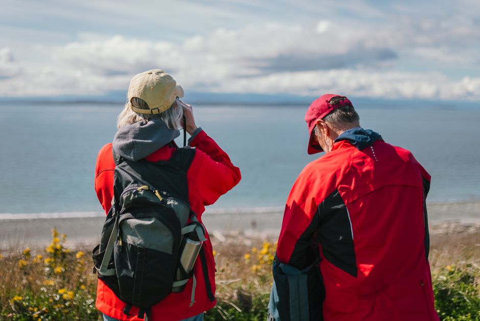 Elderly couple in red jackets enjoying seaside view, outdoors with backpacks.