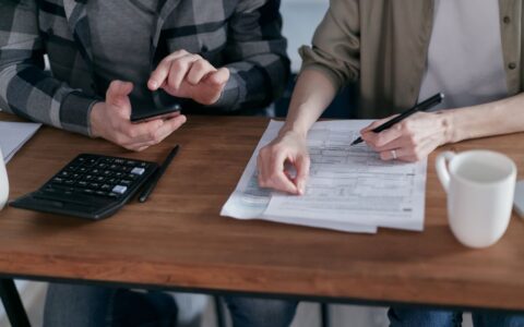 Two people working together on tax forms using a calculator at a wooden desk.