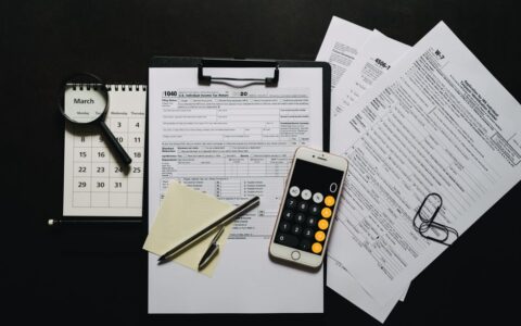 Top view of tax documents, calculator, magnifying glass, and calendar on a black surface.